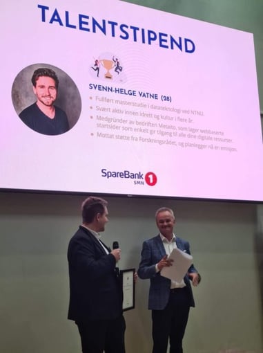 To menn med ark i hendene under en plakat det står Talentstipend på